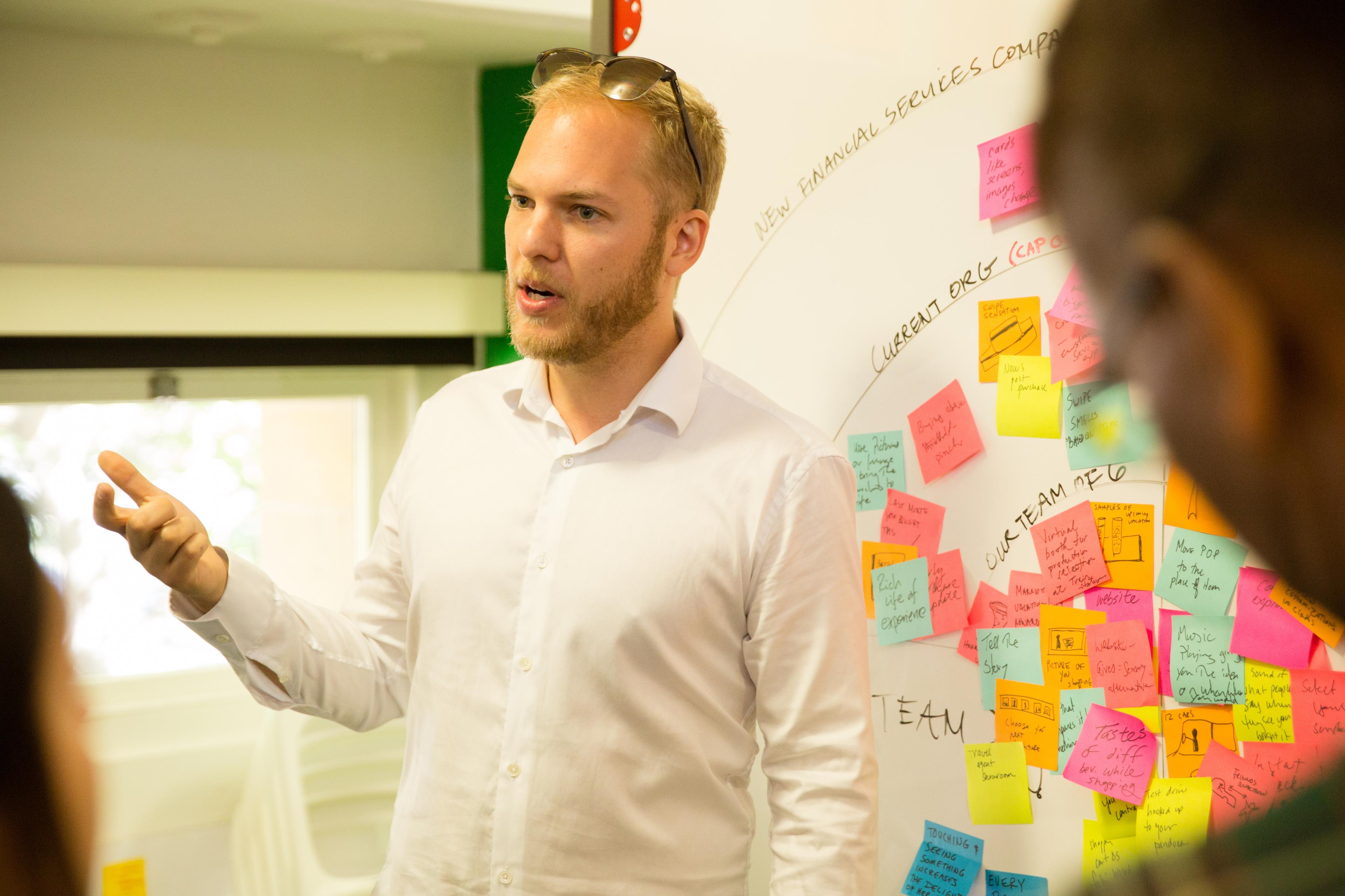 A man in a white shirt gestures while speaking in front of a whiteboard covered with colorful sticky notes during a meeting or brainstorming session.