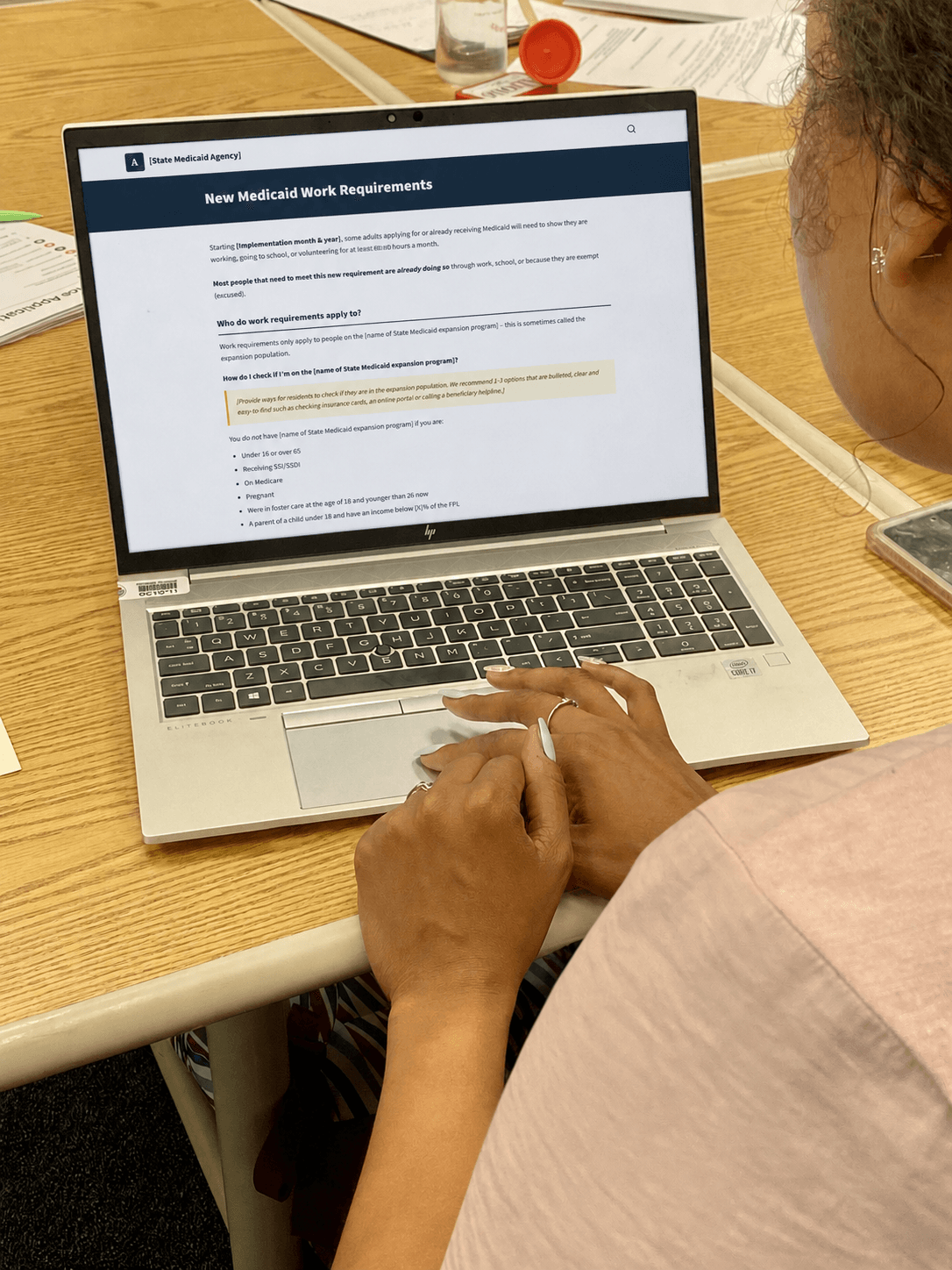 A person with painted nails reads New Medicaid Work Requirements on a laptop screen at a wooden desk, surrounded by papers and a pill bottle.