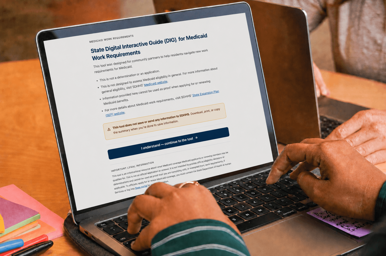 Person using a laptop displaying a webpage titled State Digital Interactive Guide (DIG) for Medicaid Work Requirements, with highlighted warnings and a blue button to continue. Colorful sticky notes and another laptop are nearby.