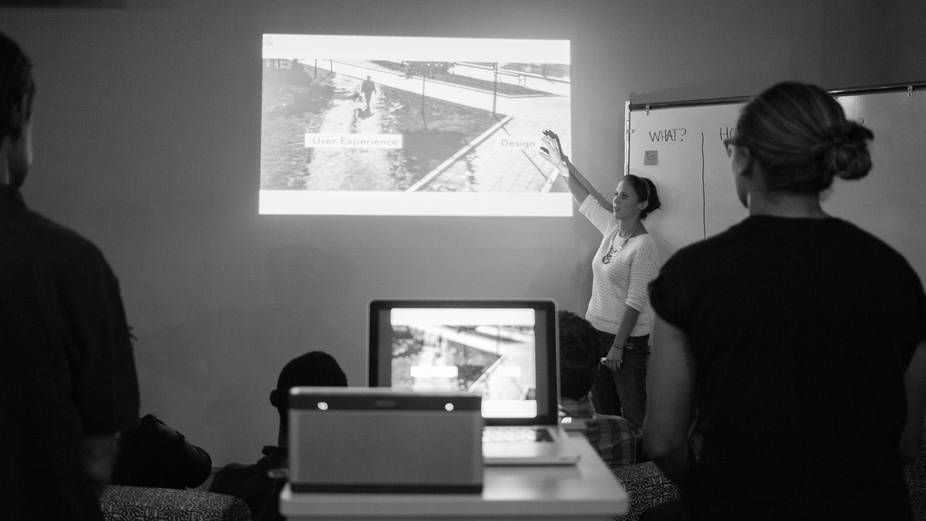 A woman stands and points at a projected presentation while several people sit and watch. A laptop and a whiteboard are visible. The slide mentions User Experience and Design..