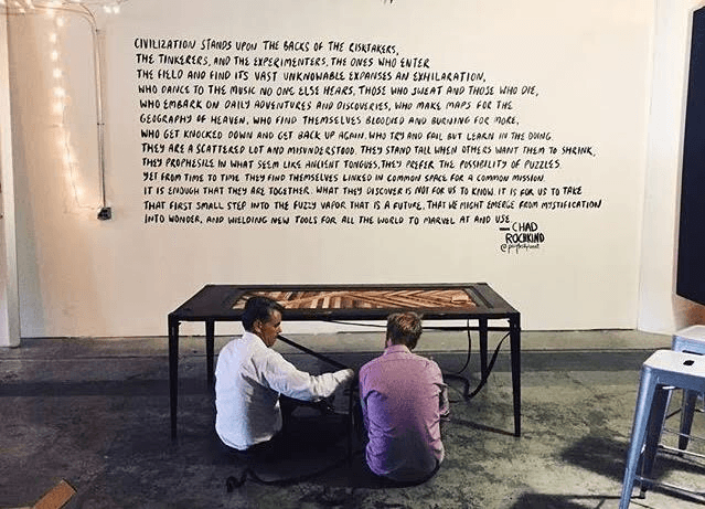 Two people sit on the floor facing each other under a table in a room with concrete floors. Behind them is a wall with a large, handwritten quote about civilization, exploration, and imagination.