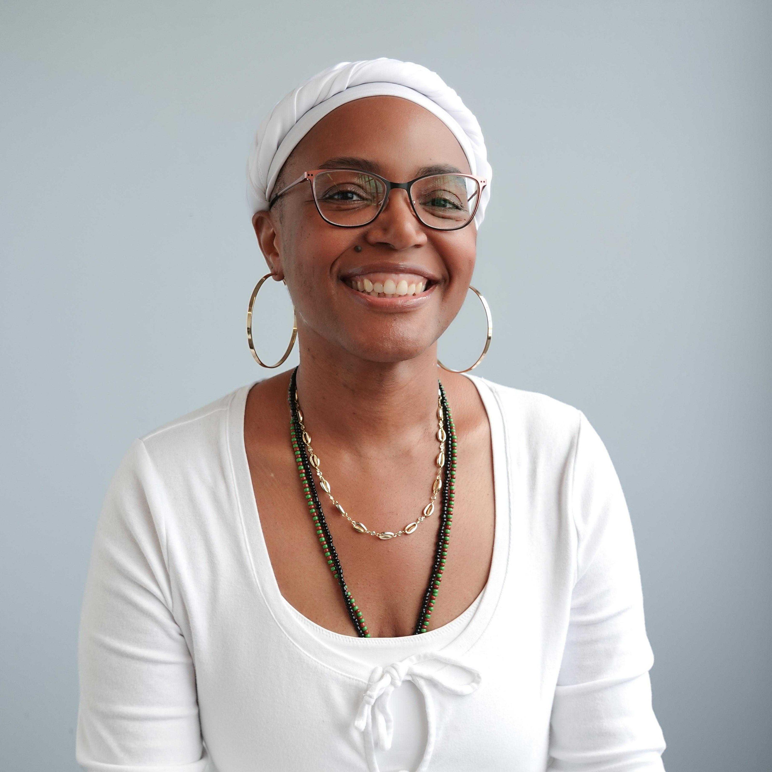 A smiling person wearing glasses, a white head wrap, large hoop earrings, layered necklaces, and a white top sits against a plain light gray background.
