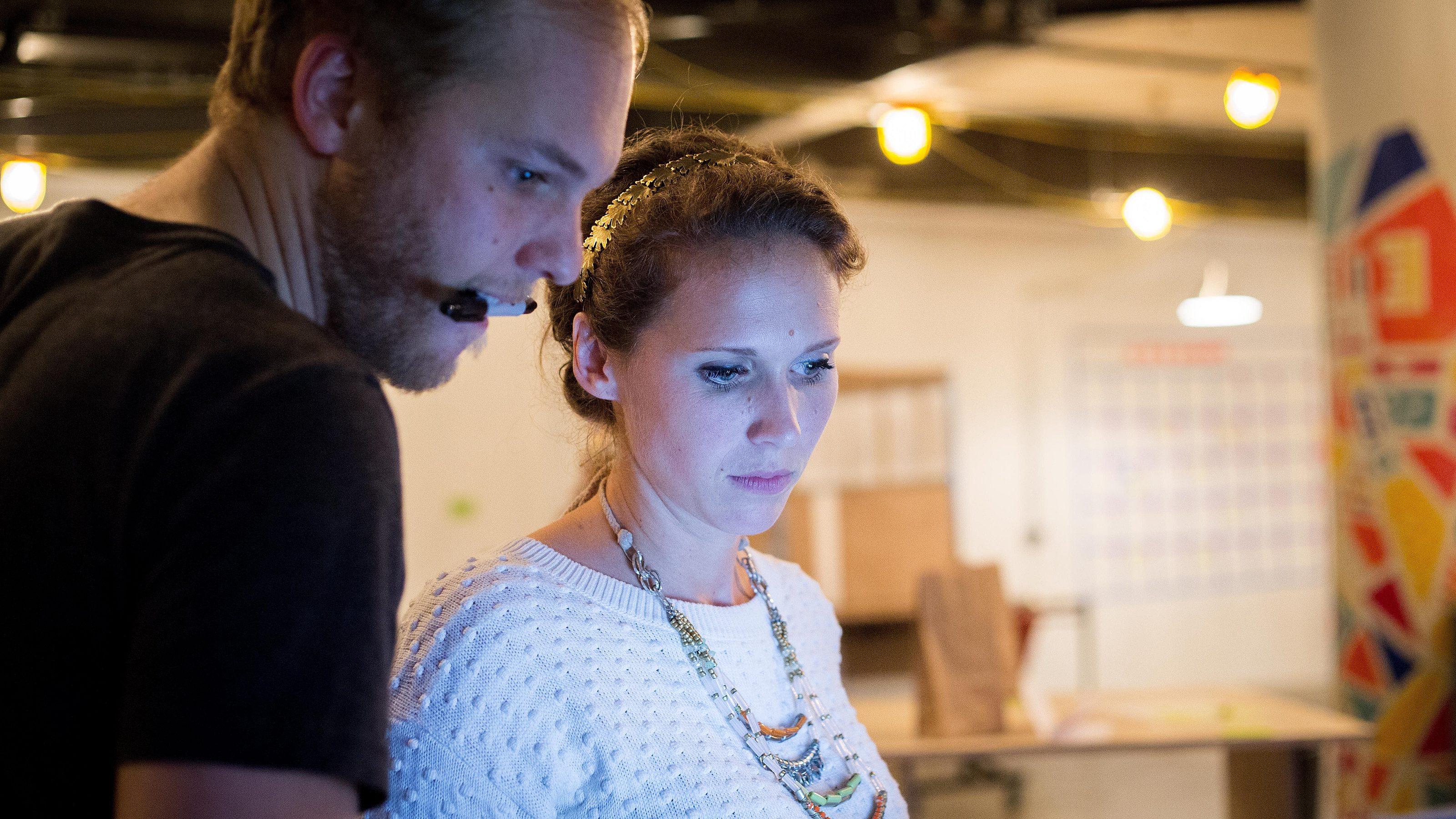 Two people stand closely together indoors, looking intently at a screen. One person wears a headset microphone; the other wears a white sweater and beaded necklace. The background is out of focus with string lights and colorful patterns.