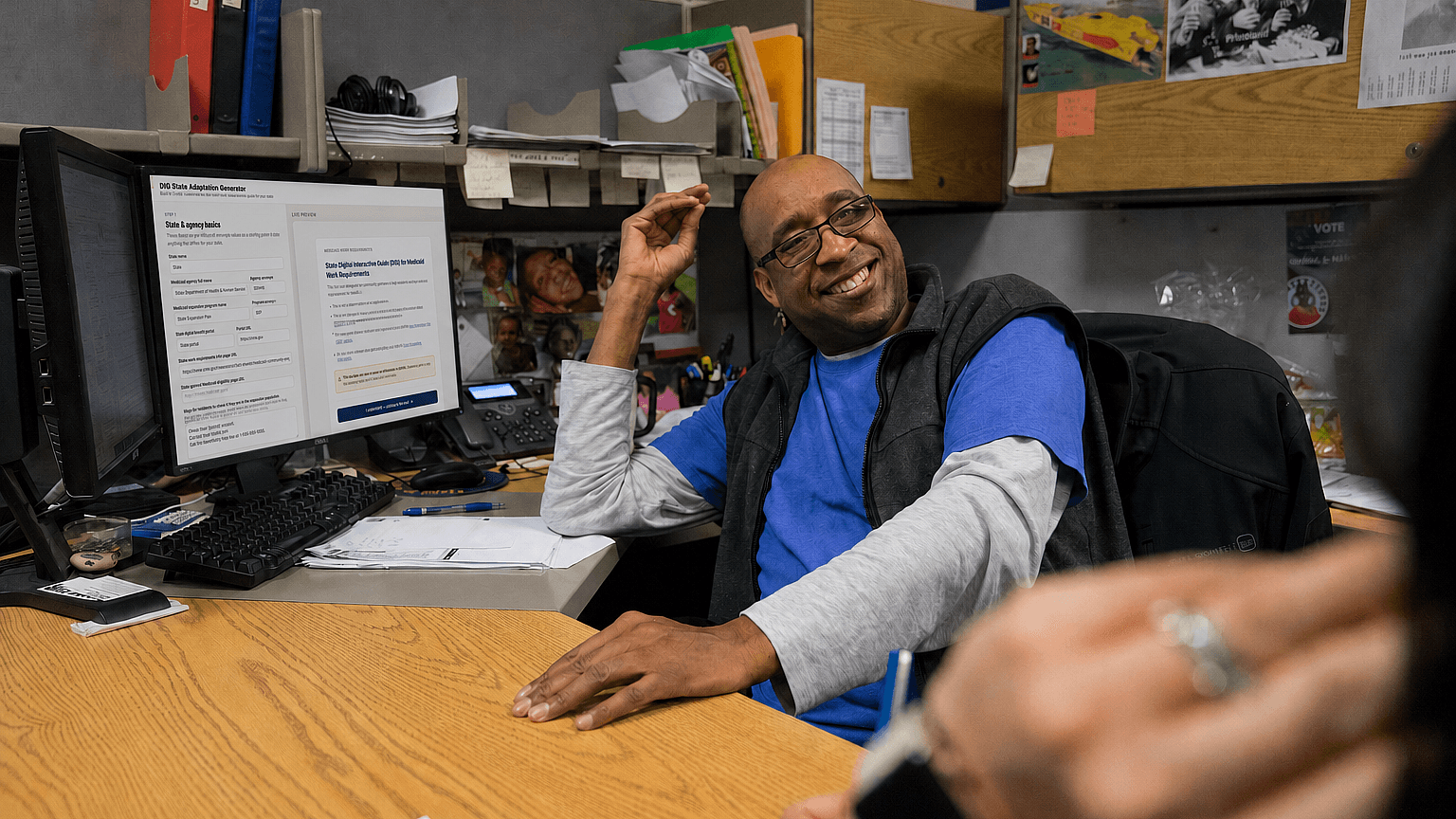 A smiling man in a blue shirt and glasses sits at a cluttered office desk, gesturing with his hand. A computer monitor displays text, and shelves with files and papers are visible in the background.