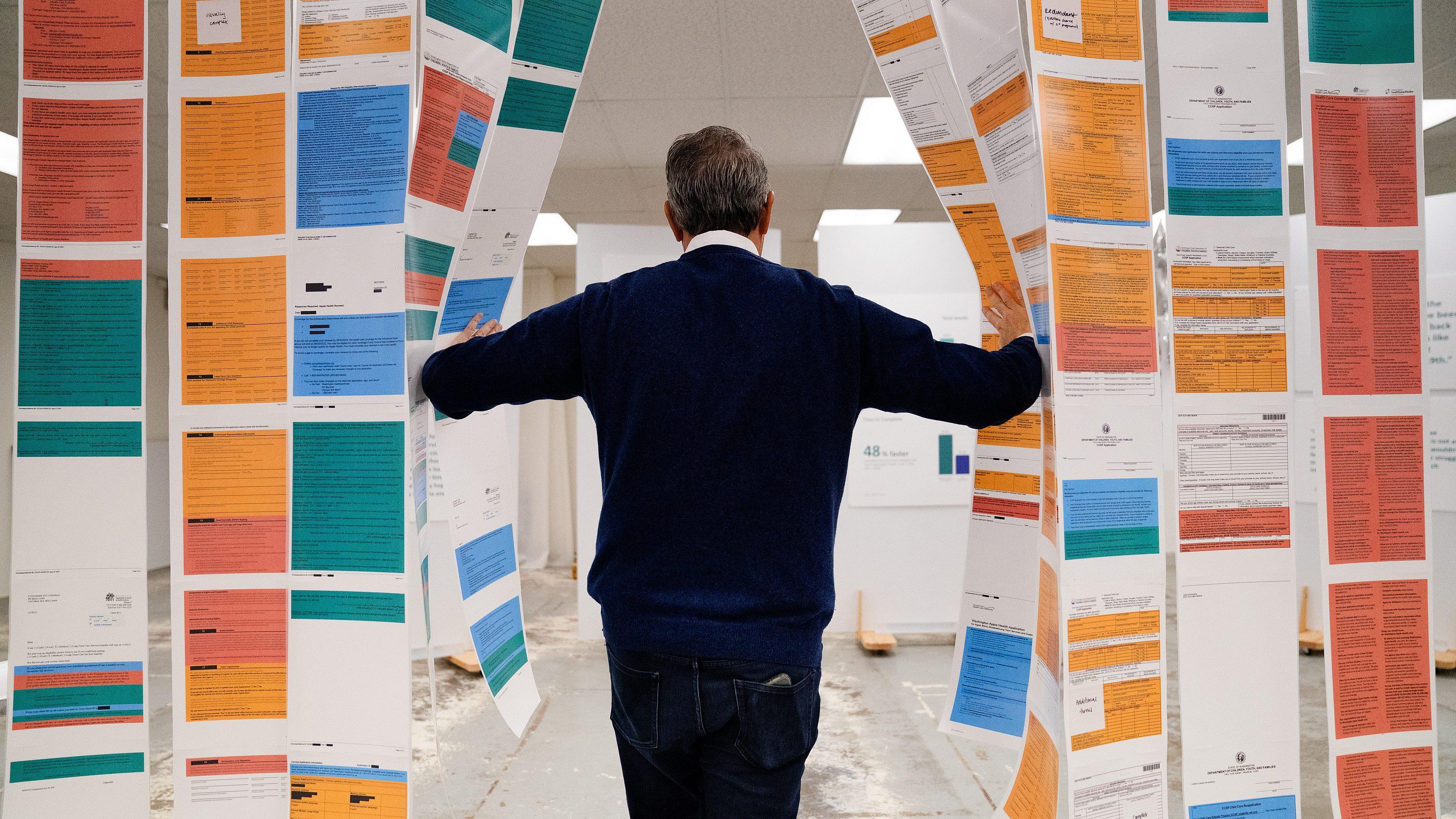 A person in a blue sweater stands with their back to the camera, walking through hanging sheets of colorful printed documents in an indoor setting.