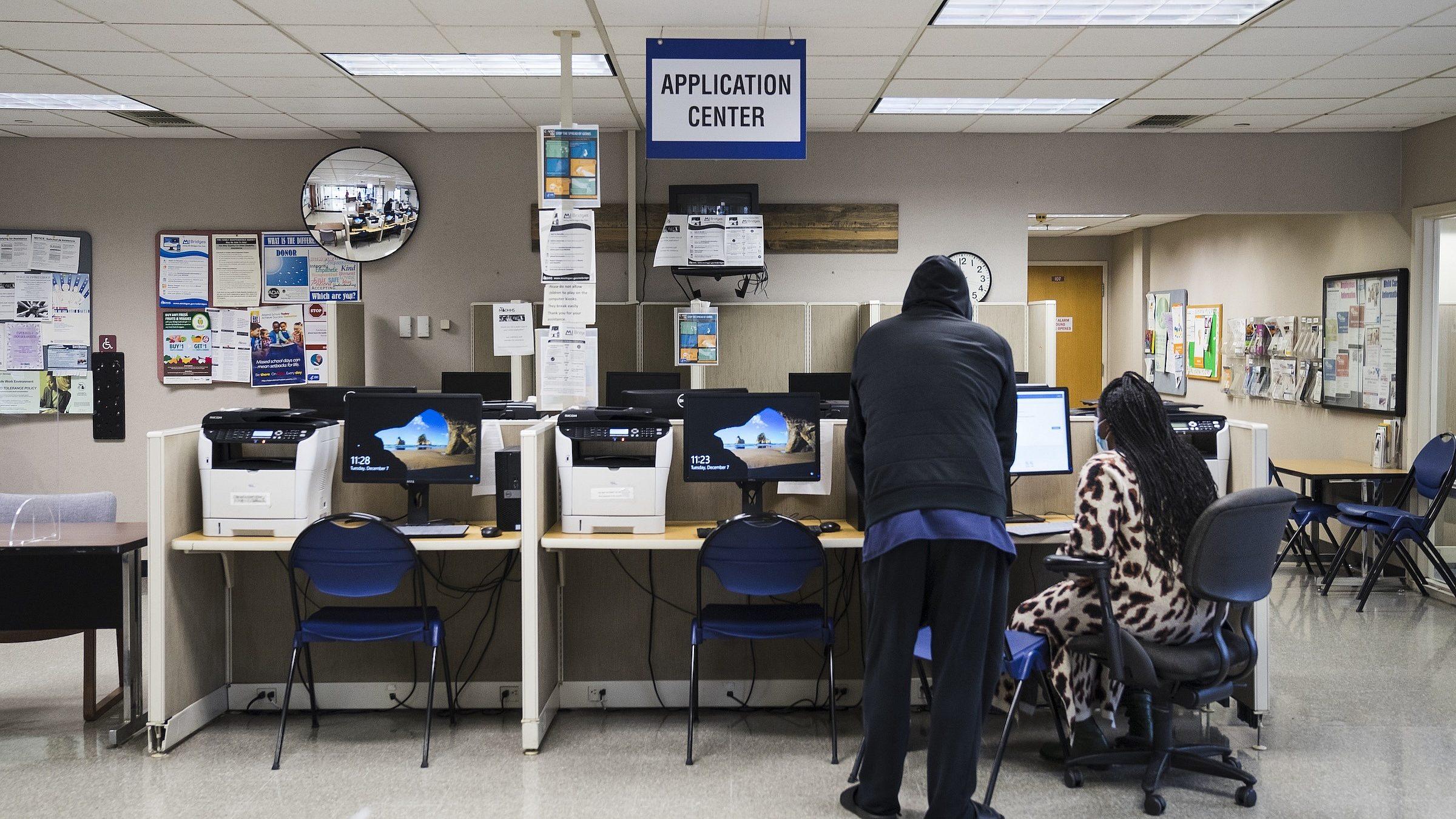 Two people use desktop computers at an application center, with several workstations, chairs, bulletin boards, and a sign overhead reading APPLICATION CENTER in a well-lit room.