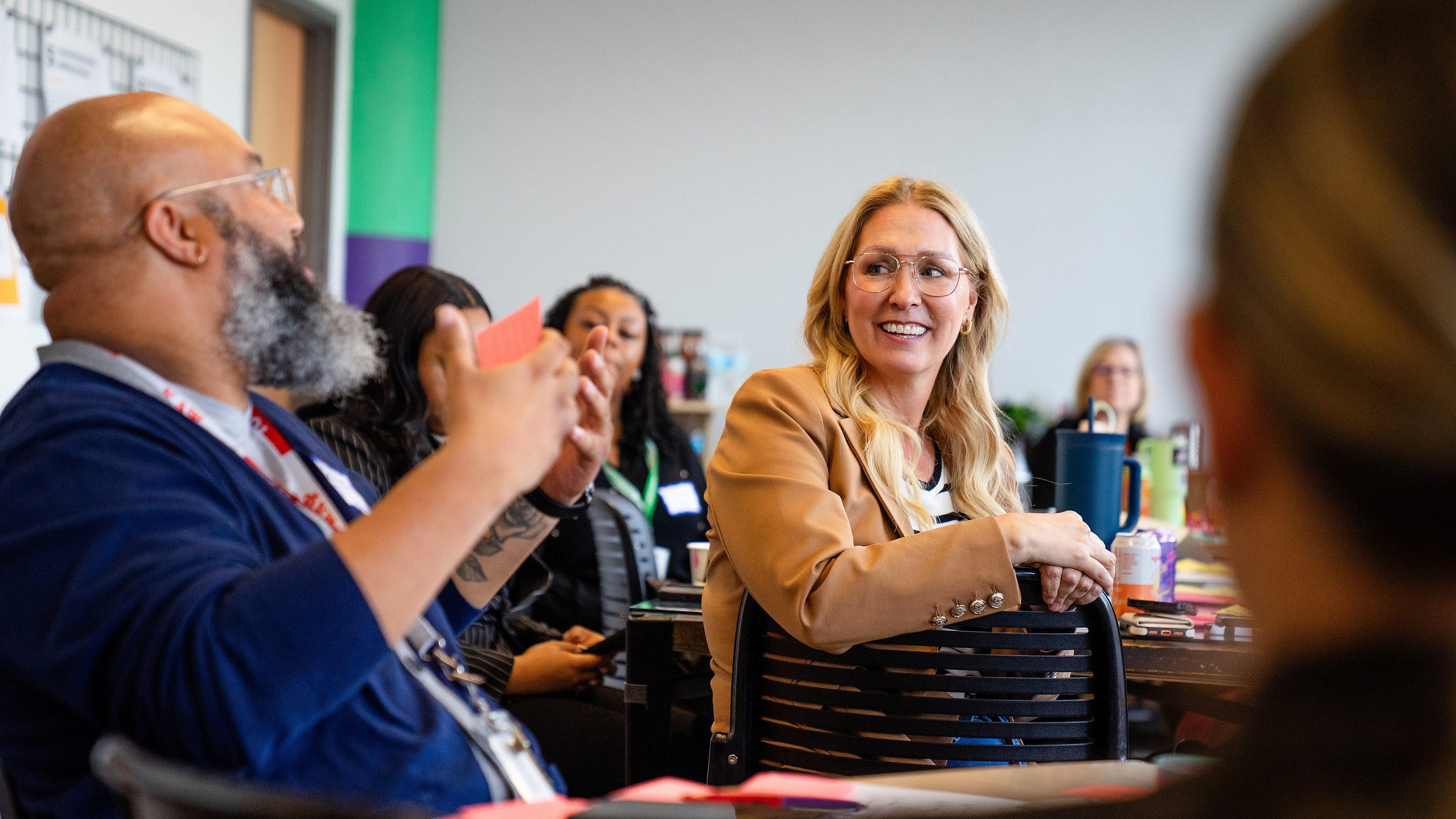 A group of people sit around a table in discussion. A woman in a tan jacket smiles and listens as a man with a beard gestures while speaking. Others in the background appear focused and engaged.