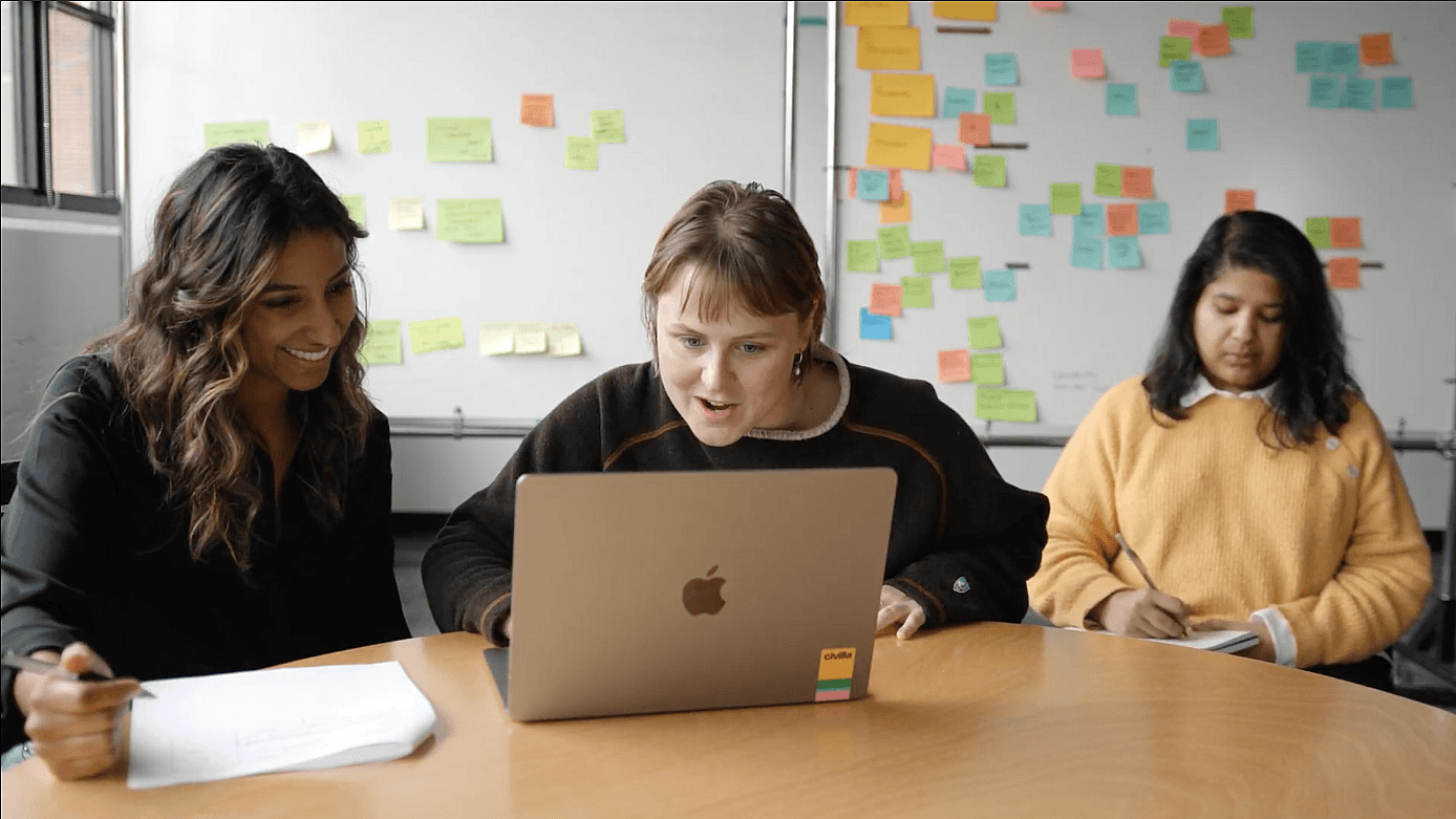 Three women sit at a round table; two smile and look at a laptop, while the third writes in a notebook. Colorful sticky notes cover the whiteboards behind them.