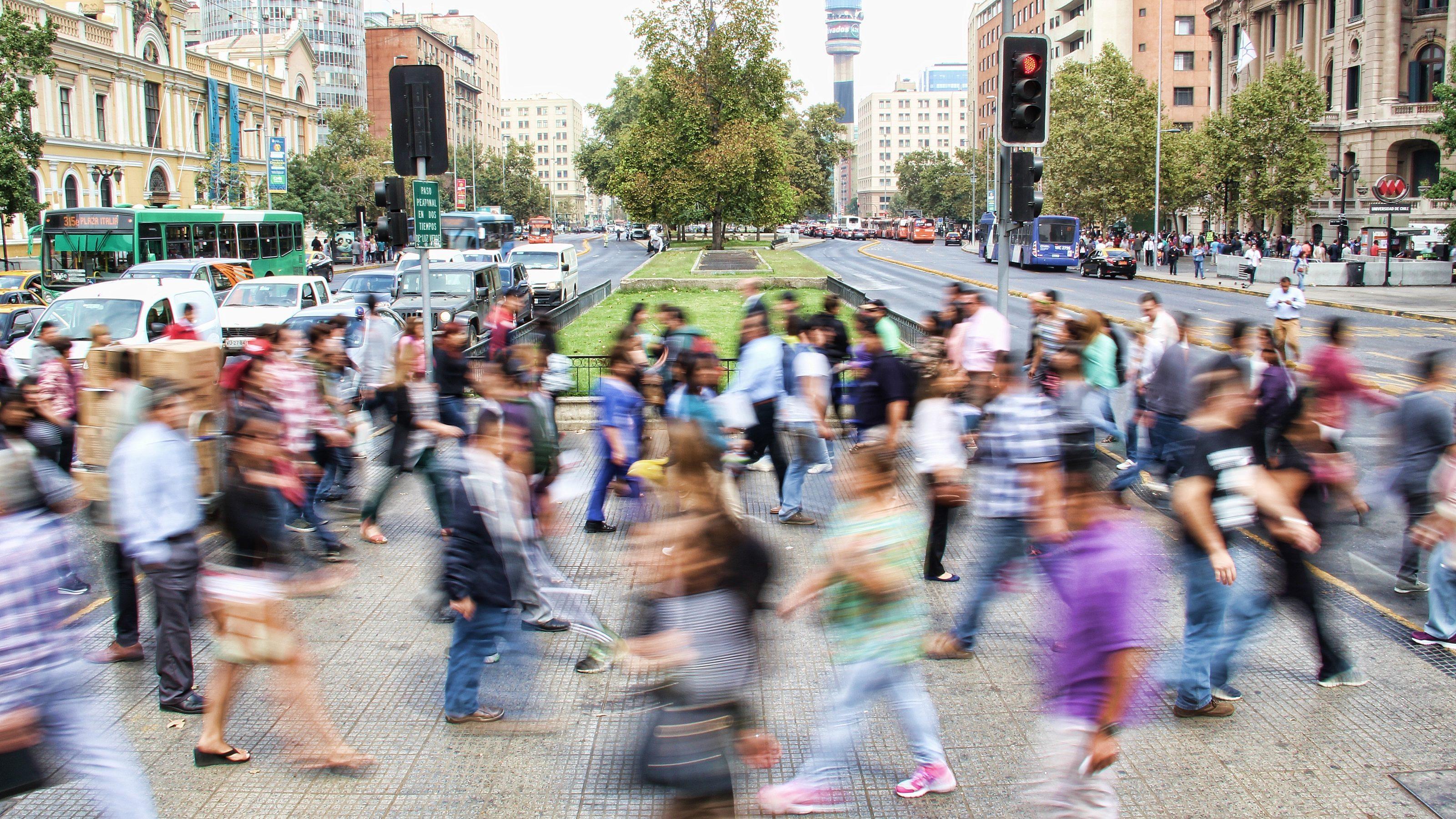 A busy city street with many people crossing at a crosswalk, their motion blurred, surrounded by buildings, cars, and traffic lights on a clear day.