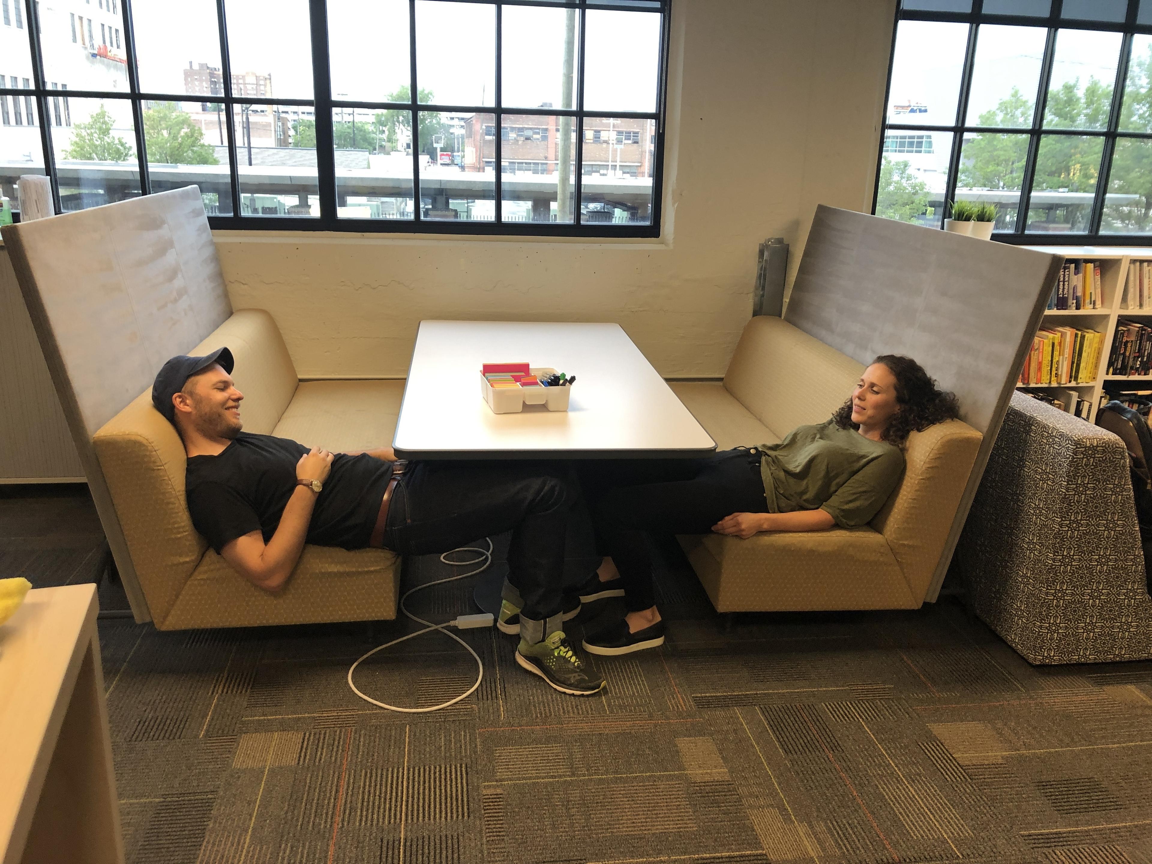 Two people relax on opposite benches of a booth in an office, with their feet resting on each others legs across the table. Office supplies sit on the table and large windows let in natural light.