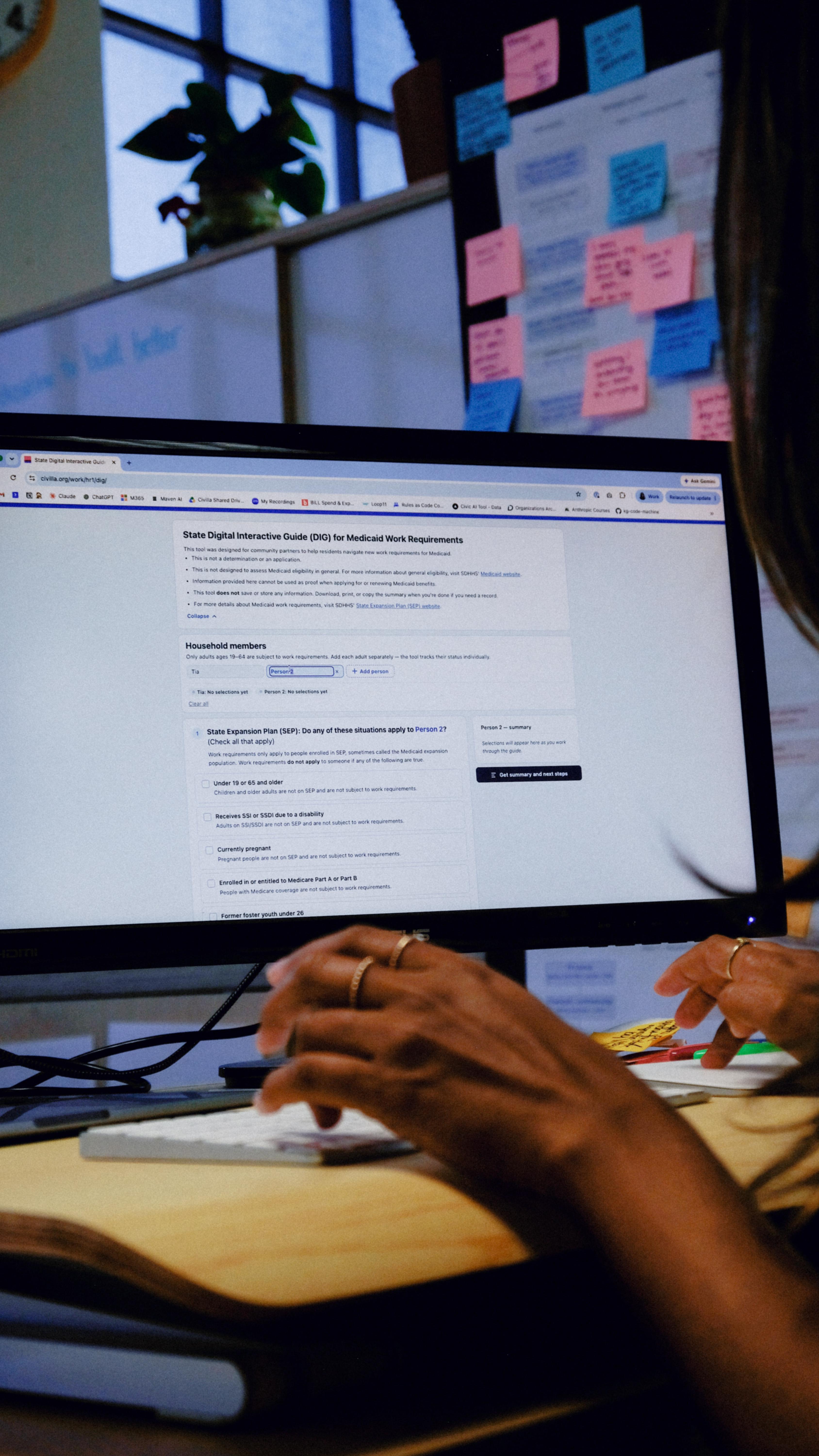 A person types on a keyboard in front of a monitor displaying a government application form, with colorful sticky notes on a wall in the background.
