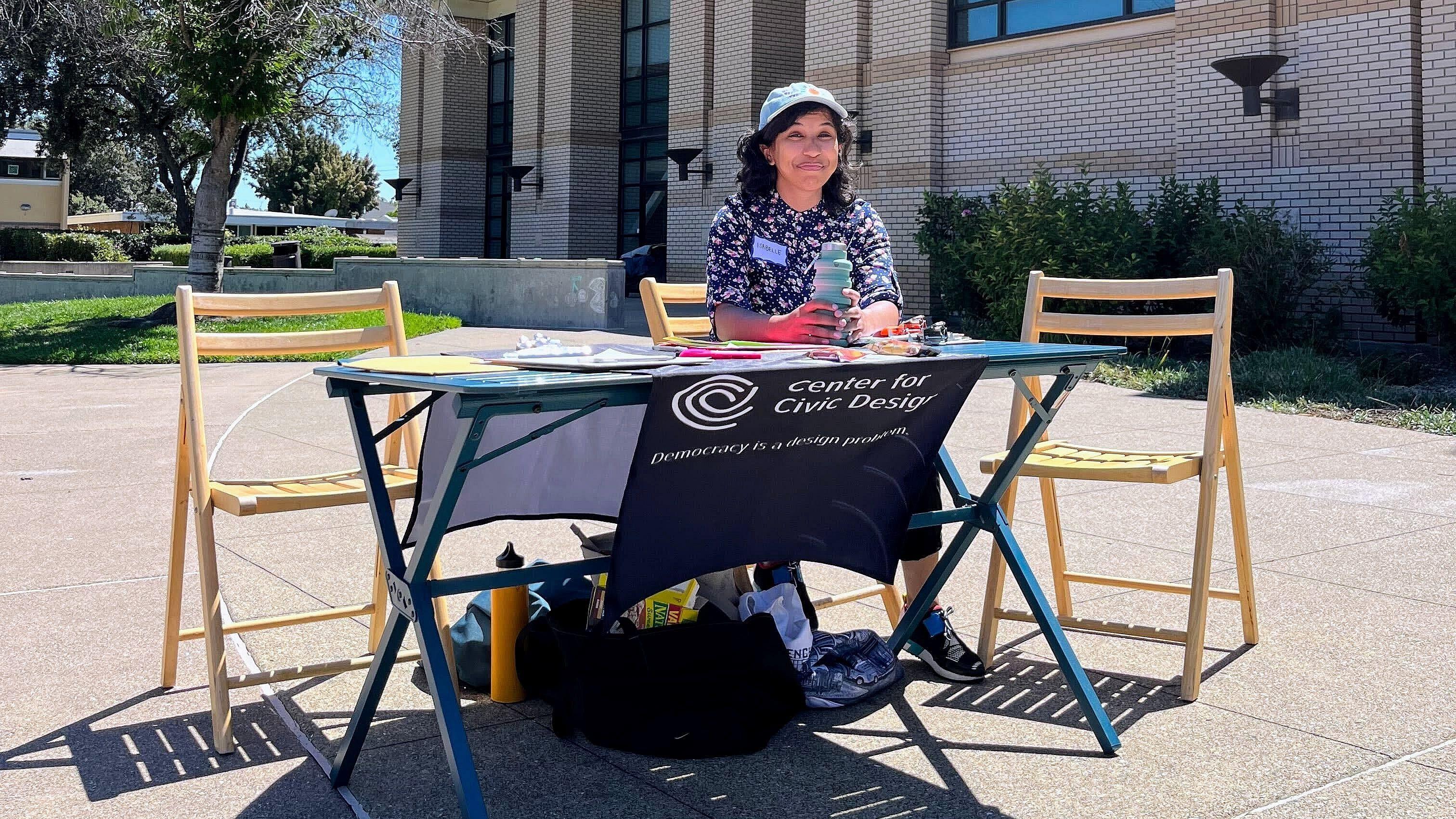 A woman sits at a table outside a building with a Center for Civic Design banner. She is wearing a hat and patterned shirt, surrounded by chairs, papers, and supplies on a sunny day.