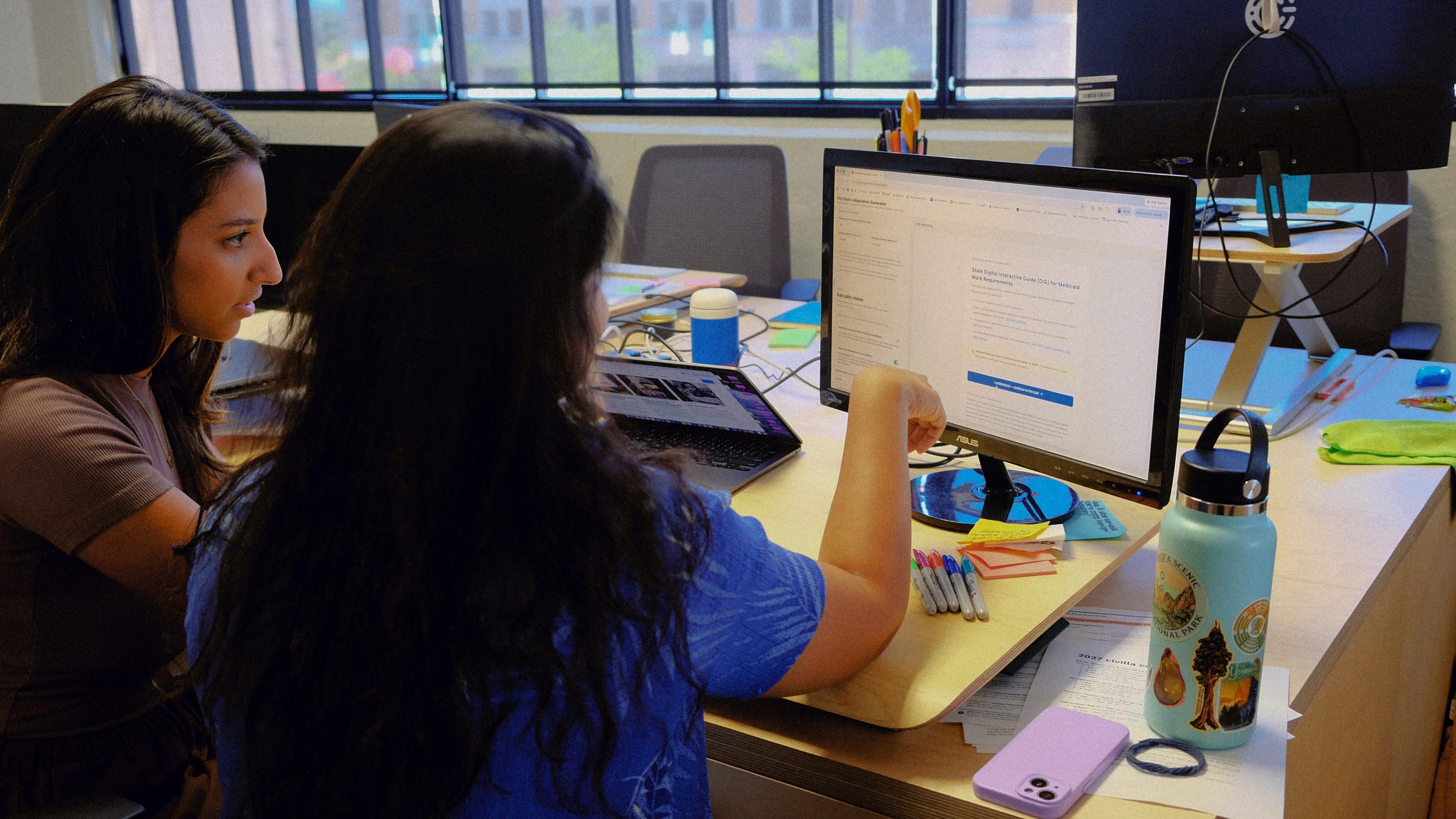 Two people sit at a desk looking at a computer screen together, with notebooks, pens, a laptop, a water bottle, and a phone on the desk in a brightly lit office with large windows.