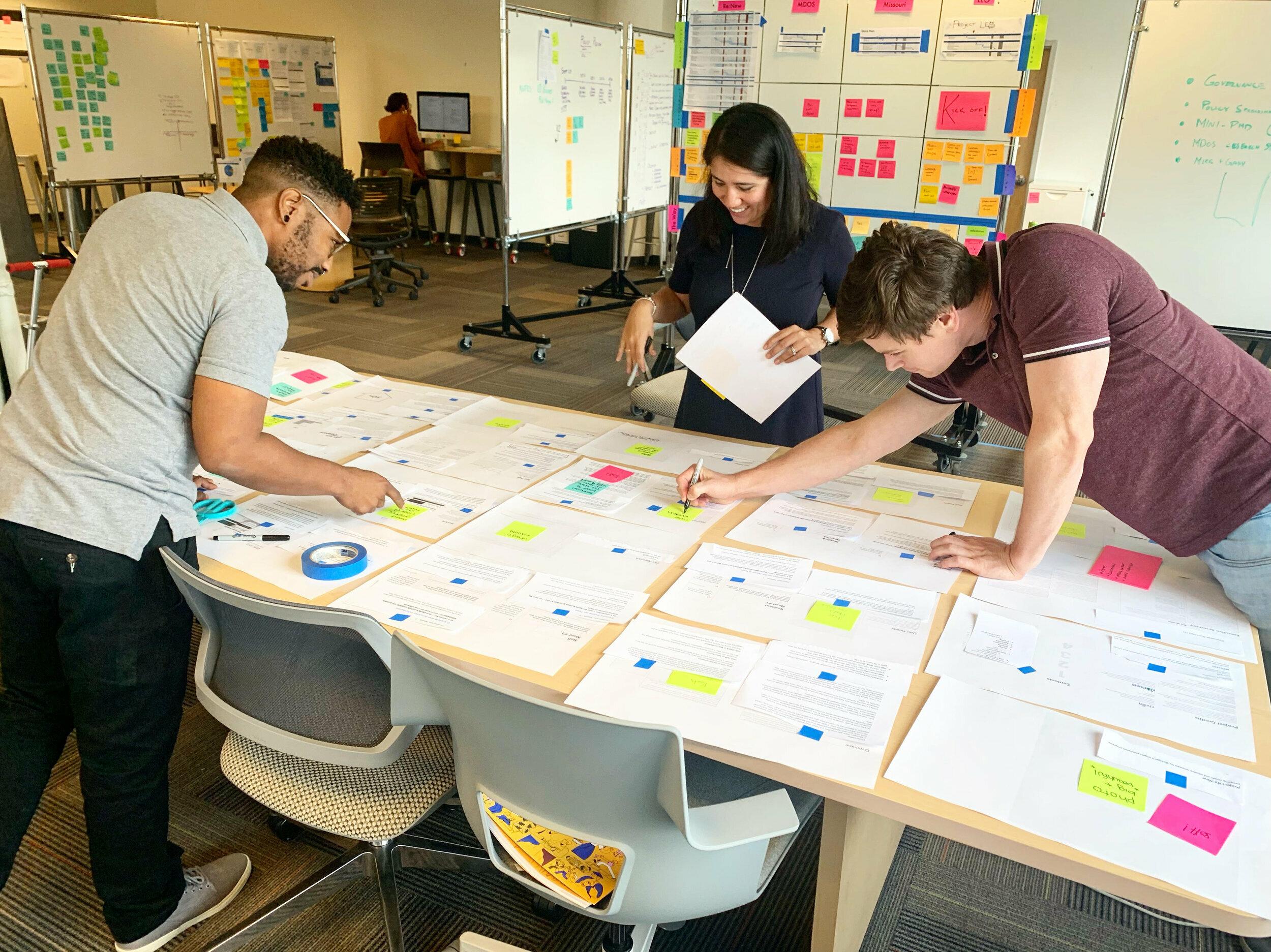 Three people are standing around a large table covered with papers and sticky notes, collaborating and making notes. Whiteboards with more notes and diagrams are visible in the background, suggesting a team brainstorming session.