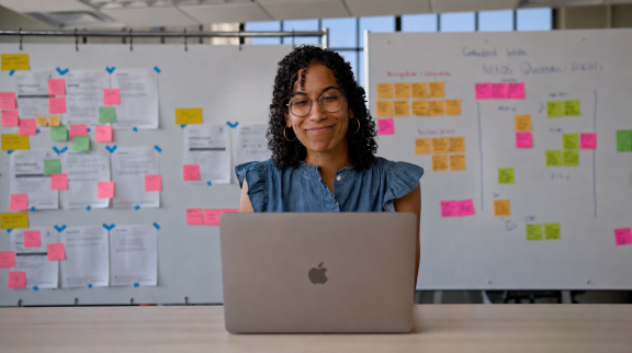 A woman with curly hair and glasses smiles while working on a laptop at a desk, with whiteboards covered in colorful sticky notes and papers in the background.