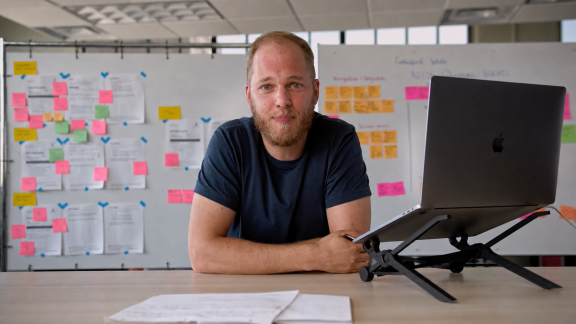A man with a beard sits at a desk with a laptop on a stand. Behind him are whiteboards covered with colorful sticky notes and papers, suggesting a collaborative or brainstorming workspace.