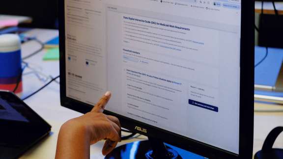 A person points at a computer monitor displaying an informational webpage, with a laptop and colorful cup visible in the background on a desk.