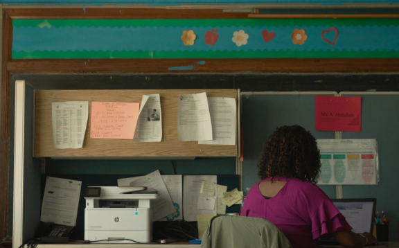 A woman with curly hair sits at a desk, working on a laptop in an office. Behind her are shelves with papers, a printer, and colorful decorations with flowers and hearts on the wall.