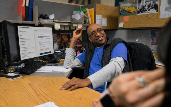 A smiling man in a blue shirt and glasses sits at a cluttered office desk, gesturing with his hand. A computer monitor displays text, and shelves with files and papers are visible in the background.