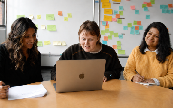 Three women sit at a table, looking at a laptop and smiling. Papers and notebooks are on the table, and colorful sticky notes cover the whiteboard in the background, suggesting a collaborative meeting or brainstorming session.