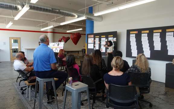 A woman stands and presents information from large boards to a seated group in Civilla's studio with industrial decor and visible ductwork. The audience watches attentively.