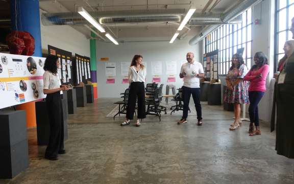 A group of six people stands in a brightly lit room in Civilla's studio  with large windows, listening to a woman presenting next to display boards with images and text. Empty chairs and posters are visible in the background.