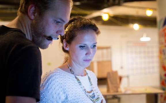 Two people stand closely together indoors, looking intently at a screen. One person wears a headset microphone; the other wears a white sweater and beaded necklace. The background is out of focus with string lights and colorful patterns.