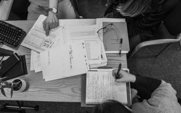 Black and white image of three people at a desk covered with papers, notebooks, and a keyboard. One person points at a document while another writes notes. A pair of glasses and a small plant are also visible on the desk.