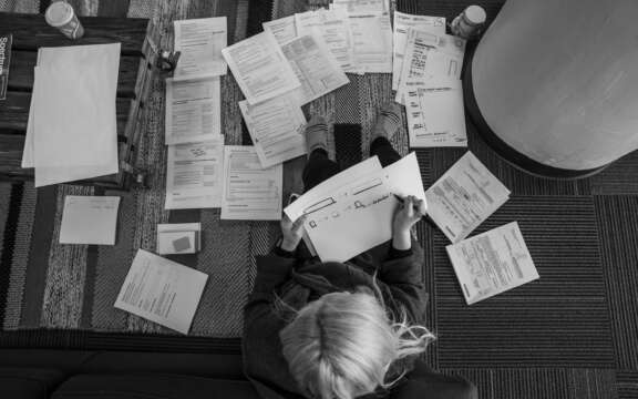 A person with light hair sits on the floor, surrounded by scattered papers and documents, holding a sheet and working with a pen. The scene is viewed from above in black and white.