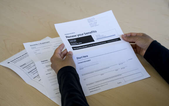 A person holds a benefits renewal form on a desk, with additional documents labeled Income + Expenses and Expenses visible nearby. The scene is well-lit and organized.
