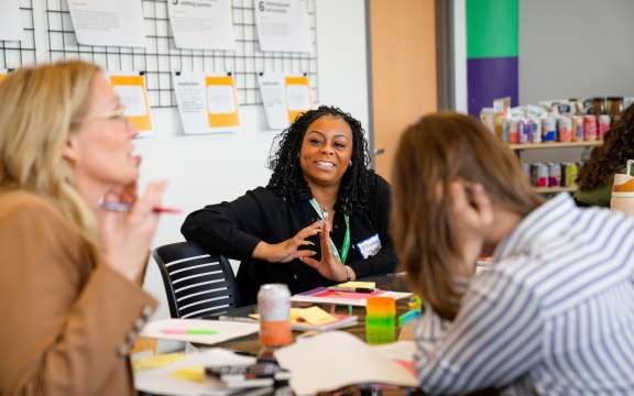 Three women sit around a table covered with papers and colorful notes, engaging in a lively discussion. One woman in the center smiles, while another gestures with her hands. Wall displays and supplies are visible in the background.
