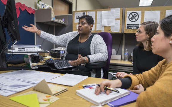 Three women sit at a desk covered with papers and notebooks, attentively listening to a fourth woman who is gesturing toward a computer monitor, holding a discussion in an office setting.