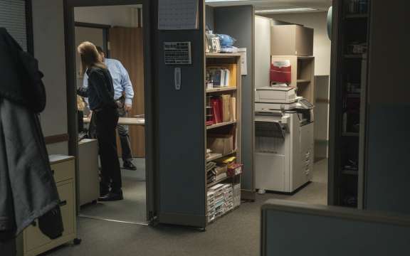 Two people stand and talk in a dimly lit office, partially visible through a doorway. The office has shelves with books, supplies, and a copy machine near the entrance. The atmosphere appears quiet and work-focused.