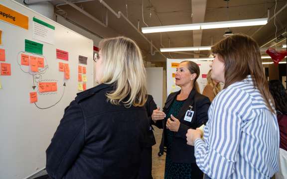 A group of women stands in front of a whiteboard covered with colorful sticky notes and diagrams, discussing ideas in a collaborative office setting.