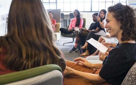 A group of people sit in a circle in a bright, modern room, engaged in conversation and holding papers, suggesting a relaxed meeting or workshop.