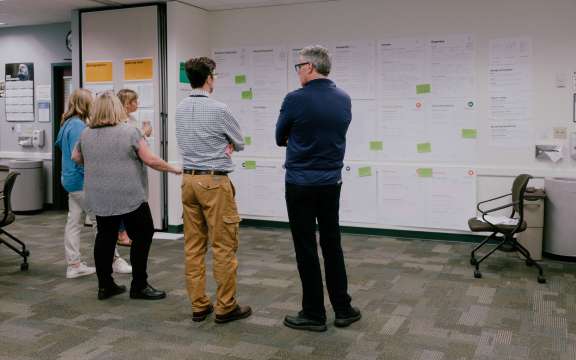 Five people stand in an office, looking at large sheets of paper with charts and notes taped to the wall, suggesting a collaborative discussion or planning session.