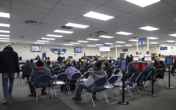 A crowded waiting area in a government office, with people seated in rows of blue chairs facing service counters and digital display screens. The environment appears busy and organized.