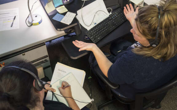 Two women wearing headsets sit at desks with computers and papers. One gestures with her hands while talking, and the other writes notes in a notebook. The workspace appears busy and organized.