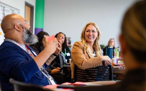 A group of people sit around a table in discussion. A woman in a tan jacket smiles and listens as a man with a beard gestures while speaking. Others in the background appear focused and engaged.