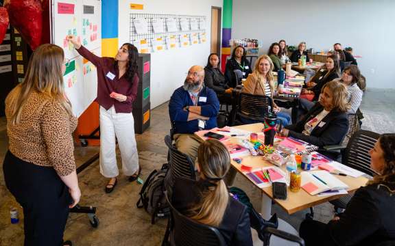 A group of adults sit around tables in a bright meeting room, listening to a woman presenting at a whiteboard covered with colorful sticky notes, as another woman stands nearby. Supplies and papers are spread on the tables.