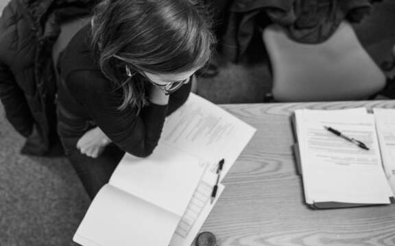 A person with glasses sits at a desk studying documents and writing in a notebook, with stacks of papers and pens nearby. The photo is in black and white and taken from above.
