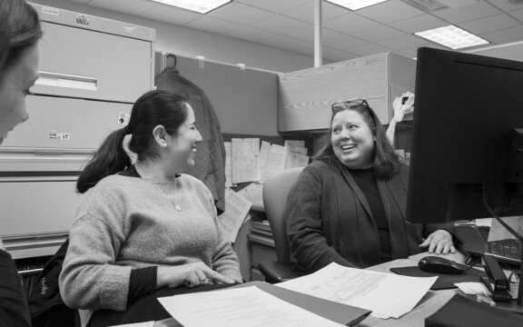 Three women sit at a desk in an office, smiling and talking to each other. Papers, a computer, and filing cabinets are visible in the workspace around them.