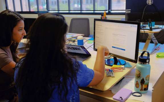 Two people sit at a desk looking at a computer screen together, with notebooks, pens, a laptop, a water bottle, and a phone on the desk in a brightly lit office with large windows.