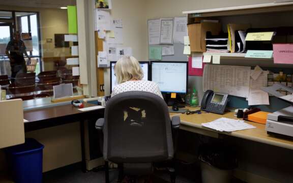 A woman sits at an office desk facing two computer monitors, surrounded by paperwork, a phone, and office supplies. The chair has a torn backrest, and the workspace appears busy and cluttered.