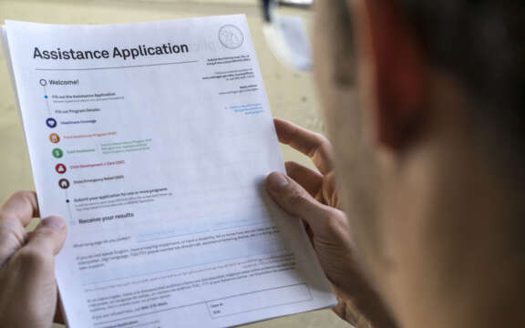 A person holding and reading an assistance application form, viewed from behind their shoulder, with the forms text and headings visible but slightly out of focus.