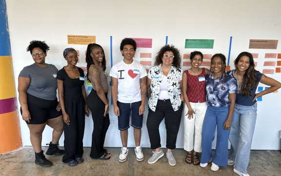 Eight people stand smiling in a row indoors in front of a wall with colorful papers and notes, dressed in casual summer clothes. The atmosphere appears friendly and relaxed.