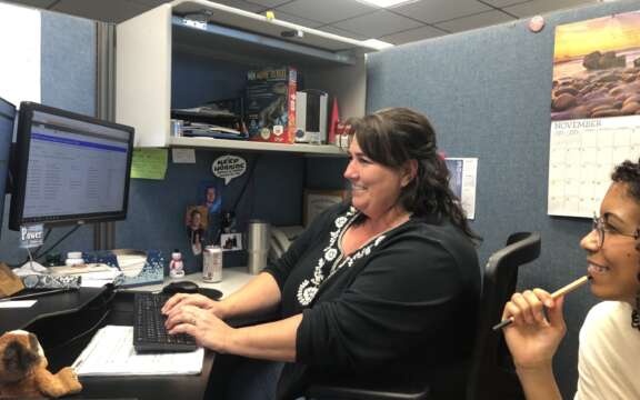 Two women sit at a desk in an office cubicle, one typing on a keyboard and smiling at a computer monitor, while the other holds a pen and looks on. Office supplies and a November calendar are visible in the background.