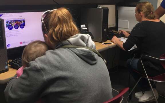 A woman holds a small child on her lap while using a computer. Another woman sits nearby at a separate computer. Both are focused on their screens in an office or computer lab setting.
