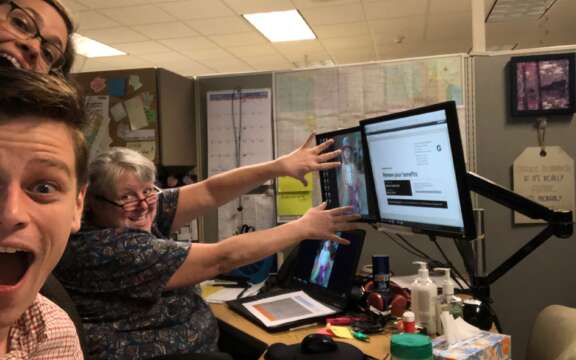 Three people in an office cubicle smile and gesture excitedly at a computer monitor displaying a webpage. The desk is cluttered with office supplies, and maps and notes are pinned on the cubicle walls.