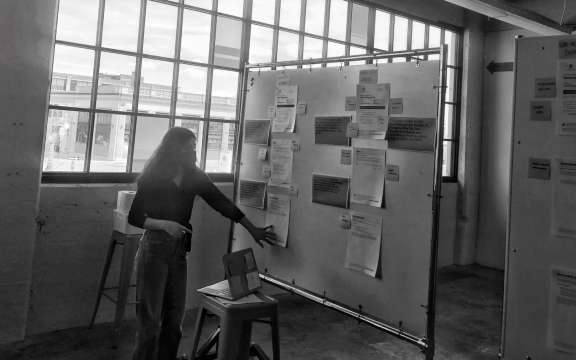 A person stands near a large board covered with papers and notes in a bright room with big windows. They are reaching toward the board, possibly organizing or reviewing the posted documents.